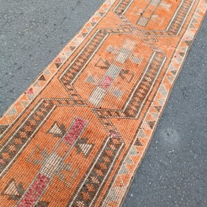 May include: A long, narrow runner rug featuring a vibrant orange background with repeating geometric patterns in shades of brown, cream, and muted red. The design includes central hexagonal motifs framed by intricate borders with triangular accents.