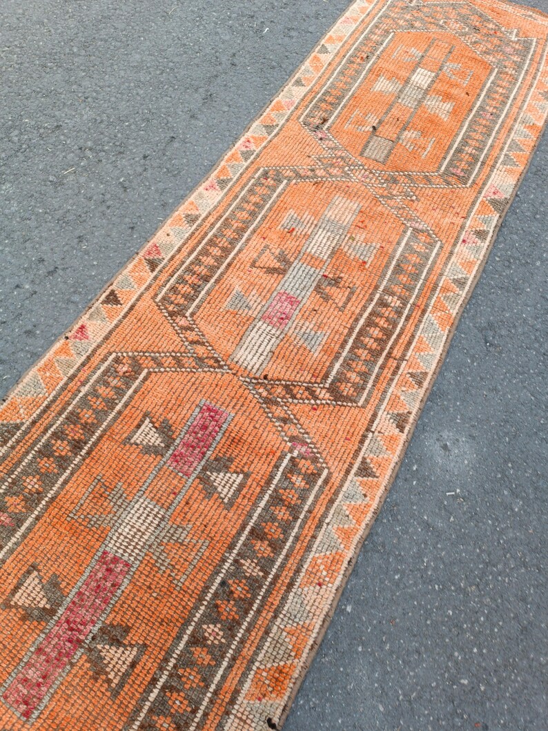May include: A long, narrow runner rug featuring a vibrant orange background with repeating geometric patterns in shades of brown, cream, and muted red. The design includes central hexagonal motifs framed by intricate borders with triangular accents.