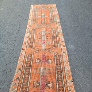 May include: A long, narrow runner rug featuring a vibrant orange background with three repeating geometric medallions in shades of brown, cream, and muted red. The border displays a repeating triangular pattern in brown and cream tones.