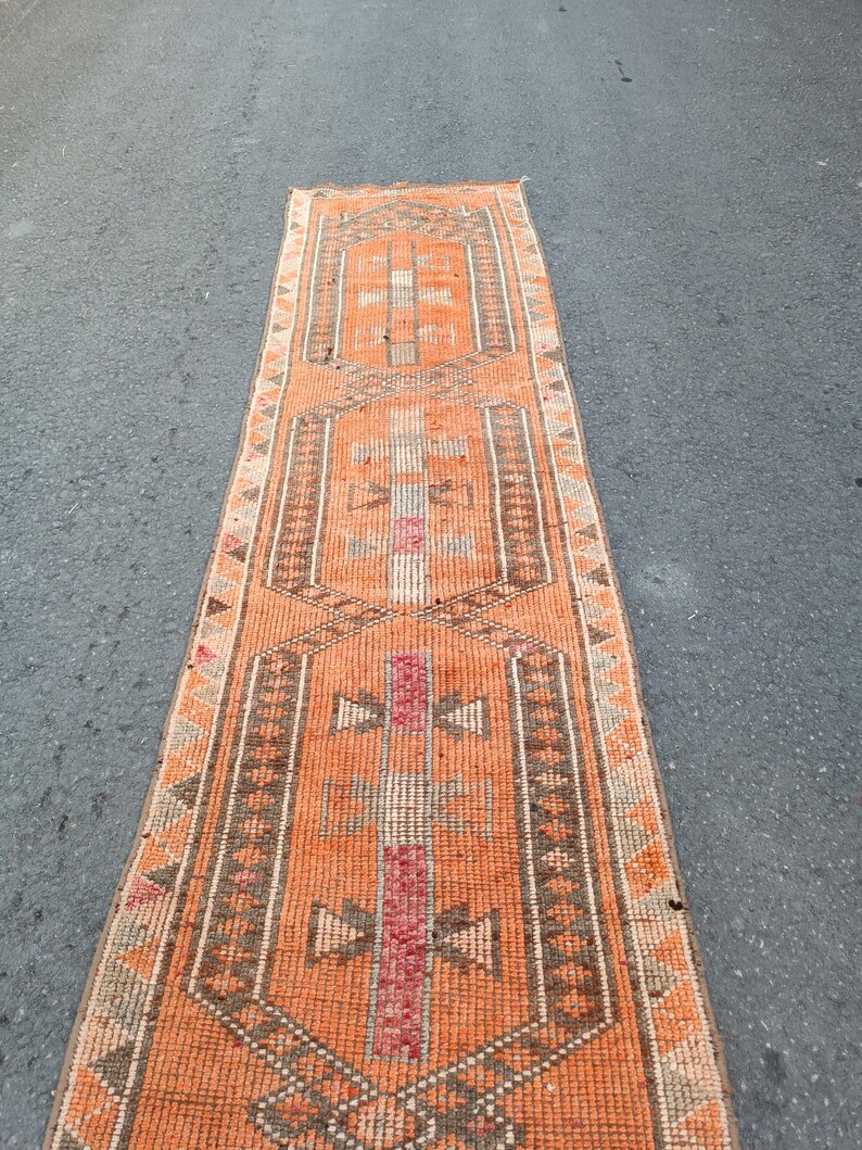 May include: A long, narrow runner rug featuring a vibrant orange background with three repeating geometric medallions in shades of brown, cream, and muted red. The border displays a repeating triangular pattern in brown and cream tones.
