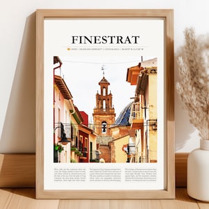 Op de afbeelding: Een ingelijste kunstdruk met een straatbeeld in Finestrat, Spanje. De afbeelding toont kleurrijke gebouwen, een kerktoren en tekst bovenaan. De print zit in een lichtgekleurde houten lijst.