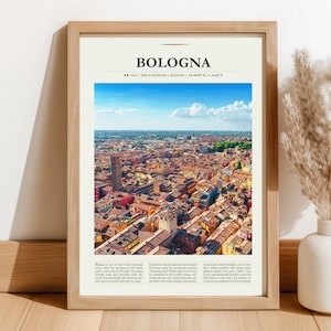 Op de afbeelding: Een ingelijste print met een luchtfoto van Bologna, Italië, met de naam van de stad bovenaan. De print toont een gedetailleerd stadsbeeld met rode pannendaken en een hoge toren tegen een blauwe lucht. De lijst is lichtbruin.