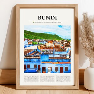 May include: A framed print featuring a vibrant cityscape of Bundi, India, with blue buildings and a green hill in the background. The word "BUNDI" is at the top, with text below. The print is in a light brown wooden frame.