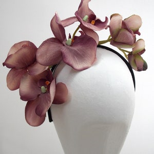 May include: A mauve orchid headband with a black elastic band. The headband is decorated with several orchid flowers.