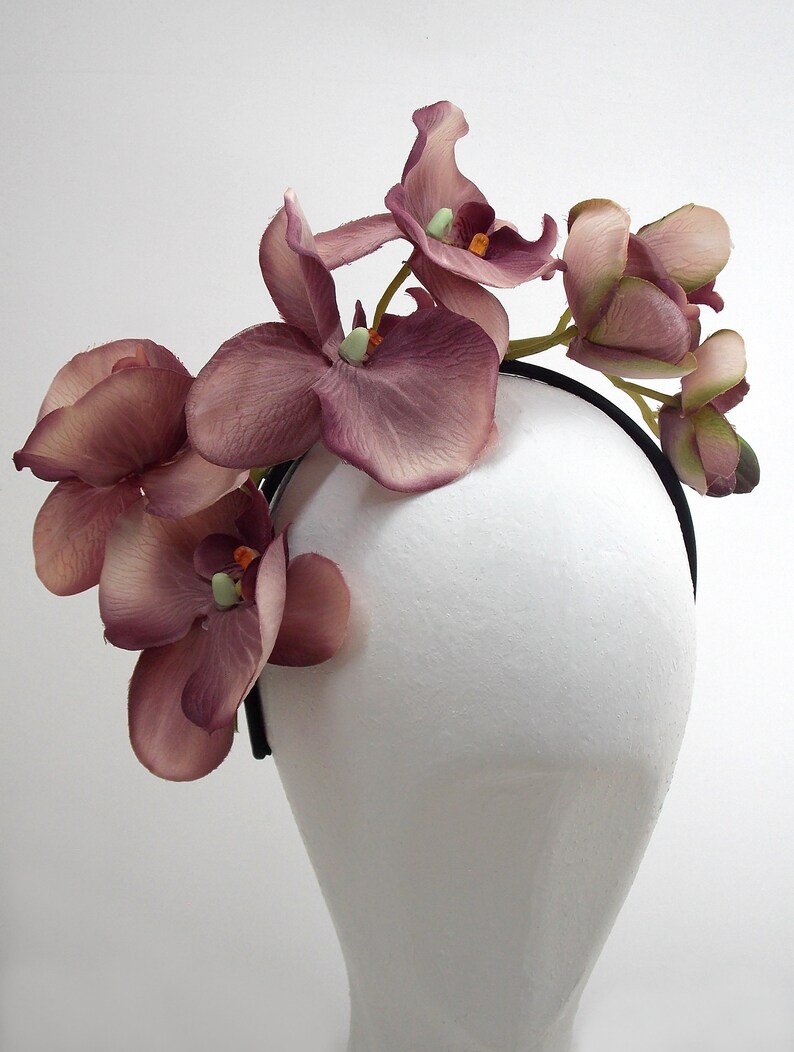 May include: A mauve orchid headband with a black elastic band. The headband is decorated with several orchid flowers.