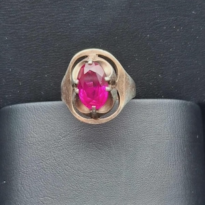 Può includere: Un anello color argento con una vistosa pietra preziosa di colore rosa fucsia acceso, dal taglio ovale e montata su quattro griffe. La fascia in metallo presenta un design liscio e arrotondato che circonda la pietra centrale.
