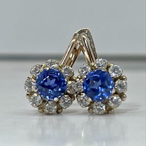 Pode incluir: Um par de brincos de ouro com fecho de alavanca, cada um apresentando uma pedra central azul redonda e facetada rodeada por um halo de pedras mais pequenas, claras, redondas e facetadas dispostas num padrão floral numa armação de ouro polido.