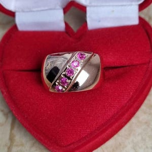 Op de afbeelding: Een gepolijste gouden ring met een diagonale zetting bezet met vijf ronde, felroze edelstenen. De ring heeft een glad, reflecterend oppervlak en wordt gepresenteerd in een rood, hartvormig fluwelen juwelendoosje met een witte binnenvoering.