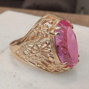 Puede incluir: Un anillo de tono dorado con una piedra preciosa rosa de forma ovalada y facetada, engastada en una banda de metal calado decorativa con intrincados patrones florales y de volutas. El engaste sujeta la gran piedra rosa con varias garras.