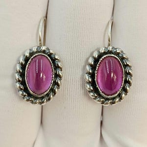 May include: A pair of silver-toned earrings. Each earring features a large, oval, magenta-colored stone set within a twisted rope-like border. The earrings have a simple hook design for pierced ears.