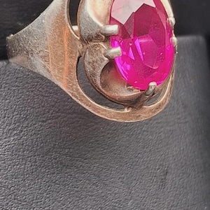 May include: A silver-toned metal ring featuring a large, oval-cut, vibrant pink gemstone set in a four-prong mounting. The band has a smooth, polished finish and a circular frame design that surrounds the central stone setting.