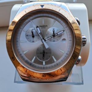 May include: A Swatch chronograph watch with a metallic gold-tone bezel and a silver-tone dial. The watch features three sub-dials, luminous hands, a date window at the four o'clock position, and a metallic link bracelet with gold and silver tones.