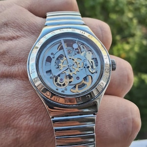 May include: A silver-toned wristwatch with a skeleton dial revealing internal gold and silver mechanical gears. The watch features a metallic expansion band and a bezel engraved with the word SWATCH repeated around the circular face.