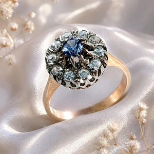 Pode incluir: Um anel de ouro com uma cravação circular em cacho, apresentando uma pedra preciosa azul-escura no centro rodeada por um halo de pedras mais pequenas de cor azul-clara. A cravação tem um design metálico rendilhado e recortado.