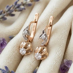May include: A pair of rose gold drop earrings featuring a hinged lever back closure. Each earring has a small round clear stone set in a textured silver-toned accent piece, with a larger round clear stone set in a rose gold flower-shaped base.