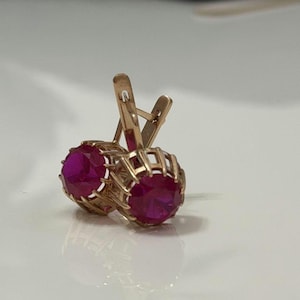 May include: A pair of gold earrings featuring round, faceted, vibrant pink stones set in a cage-like metal frame. The earrings have a hinged lever-back closure mechanism, showcasing a polished gold finish and a bright, saturated gemstone color.