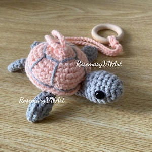 May include: A handmade crochet turtle in shades of pink and gray, with a wooden ring attached. The turtle's shell is pink with gray detailing, and the head and flippers are gray. The text "Rosemary VNArt" is visible on the image.