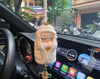 May include: A handmade car charm featuring a crocheted figure of a religious icon, adorned with a gold crown and a cross pendant with green gemstones. The charm hangs from a string of white beads, adding a touch of elegance to the car's interior.