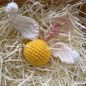 May include: A handmade crochet keychain featuring a yellow sphere with white wings and a white pom-pom. The keychain includes a pink ring and clasp. The text "Rosemary VNArt" is visible on the image.