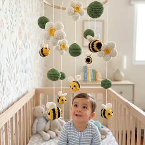 May include: A hanging mobile featuring crocheted elements suspended from a circular wooden frame. The mobile includes white flowers with yellow centers, green spheres, and small yellow and black striped bees with white wings, all attached by white strings.