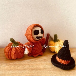 Op de afbeelding: Gehaakte Halloween-figuren, waaronder een oranje geklede Grim Reaper met een zeis, een pompoen en een heksenhoed. De Reaper heeft een wit gezicht met zwarte ogen en een gestikte mond. De heksenhoed is zwart met een oranje band.