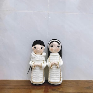 May include: Two handmade crochet dolls, one with black hair and a white robe, the other with black hair and a white headband, both in white robes with gold trim. The dolls have light skin and are standing side by side.