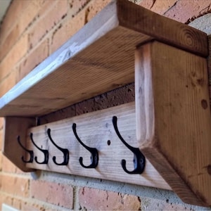 May include: A rustic wooden shelf with five black metal coat hooks. The shelf is made of light-colored wood and is mounted on a brick wall. The hooks are evenly spaced along the bottom of the shelf.