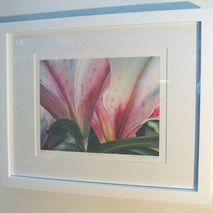 May include: A framed floral print featuring a close-up of pink and white lily petals with green leaves. The artwork is displayed in a white frame with a white mat, mounted on a light-colored wall.