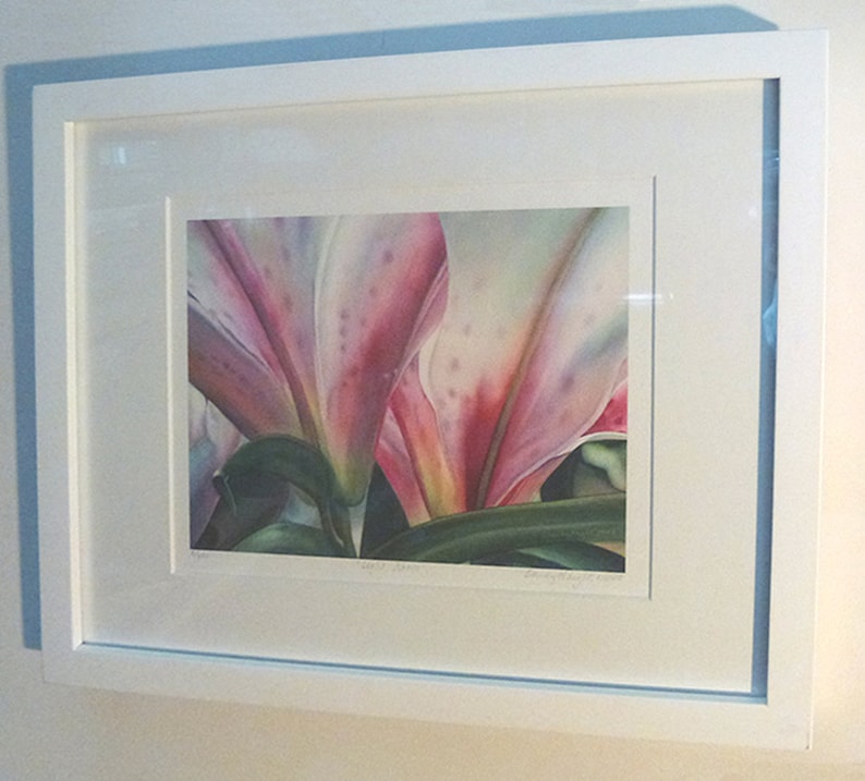 May include: A framed floral print featuring a close-up of pink and white lily petals with green leaves. The artwork is displayed in a white frame with a white mat, mounted on a light-colored wall.
