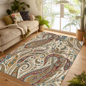 May include: A rectangular area rug with a cream background and a large paisley pattern in shades of red, blue, and gold. The rug is placed on a hardwood floor in a living room setting, with a sofa and plants visible.