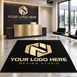 May include: A rectangular black floor rug featuring a large gold hexagonal logo and the text "YOUR LOGO HERE DESIGN STUDIO" in gold lettering. The rug is positioned on a concrete floor in front of a reception desk with a matching logo.
