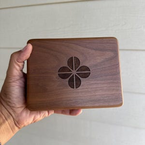 May include: A rectangular dark brown wooden box with a smooth, polished finish and a visible natural wood grain. Centered on the top surface is a laser-engraved four-petal flower design, featuring a dark, matte, and textured finish.