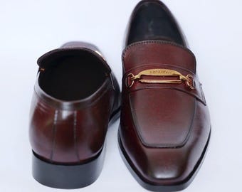 Könnte beinhalten: Ein Paar burgunderfarbene Lederschuhe mit einer goldenen Schnalle. Die Schuhe haben eine spitze Zehenpartie und einen niedrigen Absatz.