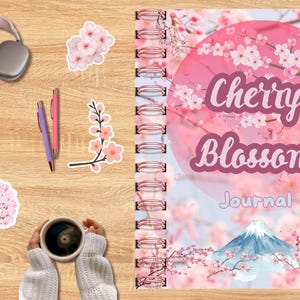 May include: A wooden surface displays a Cherry Blossom Journal, stickers, pens, headphones, and a cup of coffee. The journal cover features a pink floral design with the words "Cherry Blossom Journal". Stickers include cherry blossoms and a branch.