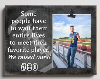 May include: A rectangular wooden photo display board with a dark gray distressed finish. The text reads: Some people have to wait their entire lives to meet their favorite player. We raised ours! #80. A photo of a football player is clipped to the board.