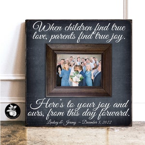 May include: A black chalkboard sign with a white frame and a photo of a wedding party. The sign reads "When children find true love, parents find true joy. Here's to your joy and ours, from this day forward. Lindsey & Jeremy - December 3, 2022"