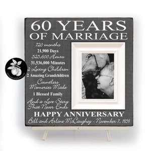 May include: A gray wooden picture frame with a white mat and a black and white photo of an older couple. The frame has a white inscription that reads "60 Years of Marriage" and includes details like "720 months", "21,900 Days", "525,600 Hours", "31,536,000 Minutes", "2 Loving Children", "2 Amazing Grandchildren", "Countless Memories Made", "1 Blessed Family", "And a Love Story That Never Ends", "Happy Anniversary", "Bill and Arline McCaughey - November 7, 1959".