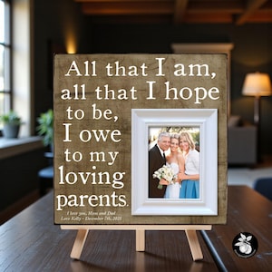 May include: A wooden framed photo with a white border, resting on a small easel. The frame has the text "All that I am, all that I hope to be, I owe to my loving parents." A photo of three people is inside the frame. The text "I love you, Mom and Dad Love Kelly - December 7th, 2025" is at the bottom.