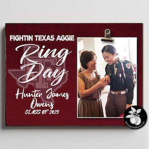 Puede incluir: Un letrero rectangular de madera color granate con el texto blanco FIGHTIN TEXAS AGGIE RING DAY, Hunter James Owens y CLASS OF 2025. Un clip de metal sostiene una fotografía de dos personas, una con vestido marrón y otra con uniforme militar oscuro.