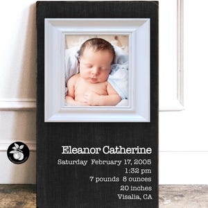 May include: A framed photograph of a newborn baby, set against a black textured background. The white frame surrounds the baby, who is sleeping peacefully. The text below reads "Eleanor Catherine", birth date, time, weight (7 pounds 8 ounces), and height (20 inches).