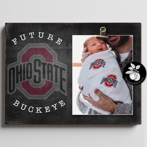 May include: A black wooden picture frame with the words "FUTURE OHIO STATE BUCKEYE" printed in white. The frame holds a photo of a baby wrapped in a white blanket with the Ohio State logo.
