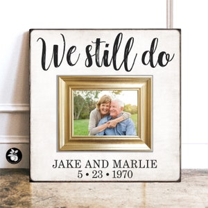 May include: A white wooden sign with a gold frame picture of an older couple. The sign says "We still do" and "JAKE AND MARLIE 5.23.1970".