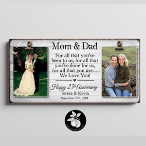 May include: A rectangular wooden sign with a white-washed finish featuring two metal clips holding photographs. The text reads: Mom & Dad, For all that you've been to us, for all that you've done for us, for all that you are... We Love You! Happy 25th Anniversary, Teresa & Kevin, November 15th, 2001.