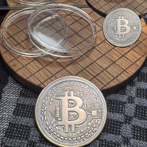 May include: A close-up of two Bitcoin coins with a gold and silver finish. The coins are on a wooden surface with clear plastic containers. The Bitcoin symbol is prominently displayed on the coins.