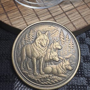 May include: A round, bronze-colored coin featuring a detailed relief of a wolf pack in a forest setting. The coin depicts four wolves, with trees and mountains in the background. The coin is on a dark, patterned surface.