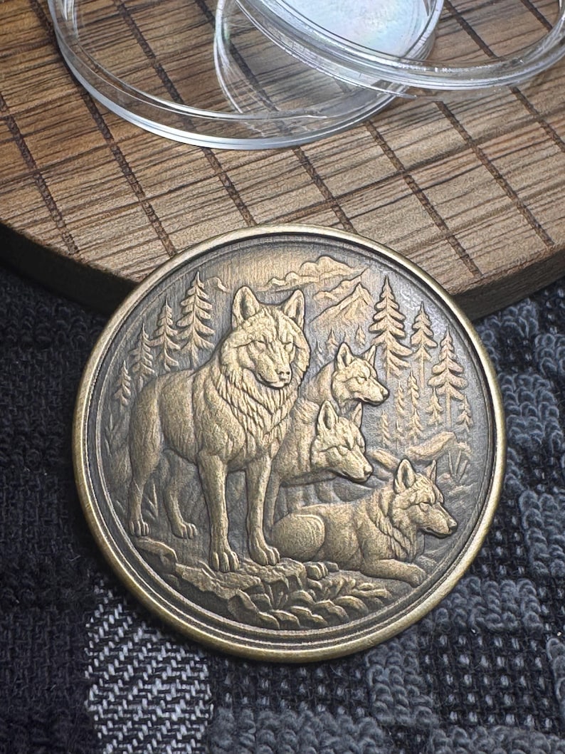 May include: A round, bronze-colored coin featuring a detailed relief of a wolf pack in a forest setting. The coin depicts four wolves, with trees and mountains in the background. The coin is on a dark, patterned surface.