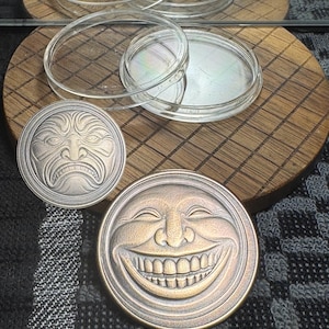 May include: Two metal coins with detailed face designs, one with a stern expression and the other with a wide grin. Clear, circular petri dishes are arranged on a wooden surface, with a reflection visible above. The background is a patterned fabric.