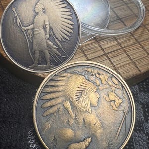 May include: Two bronze-colored coins with Native American designs. One coin features a figure holding a staff, the other depicts a profile with a wolf howling. A clear plastic case sits above the coins. The coins are on a wooden surface.