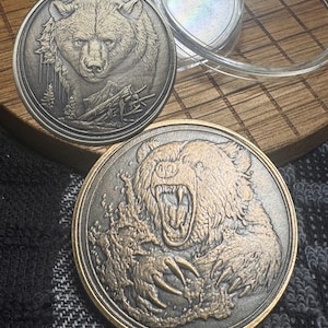 May include: Two round, bronze-toned coins featuring detailed bear imagery. One coin depicts a bear's head with a forest scene, while the other shows a roaring bear with claws extended. The coins are displayed near a clear plastic case.