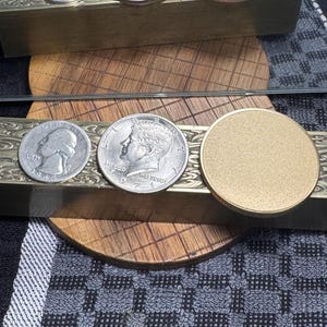 May include: Two brass rectangular boxes with decorative edges, each holding two coins and a gold-colored disc. The coins include a quarter and a half-dollar. The boxes are on a wooden surface.